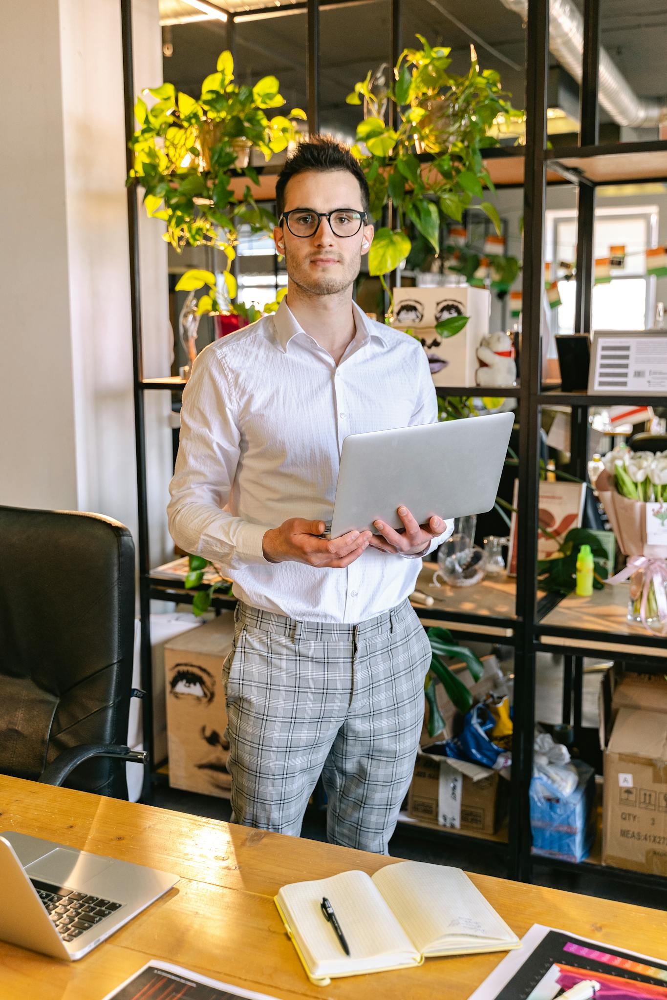 A young man holding a laptop in a contemporary office with plants and creative decor.