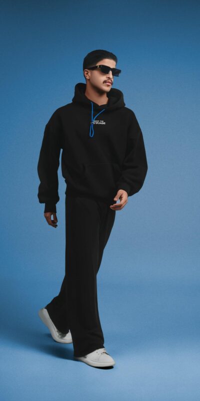 Fashionable man in black hoodie and pants walking against a blue backdrop.