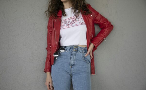 Fashionable woman in red jacket and jeans posing against a gray wall, showcasing casual style.
